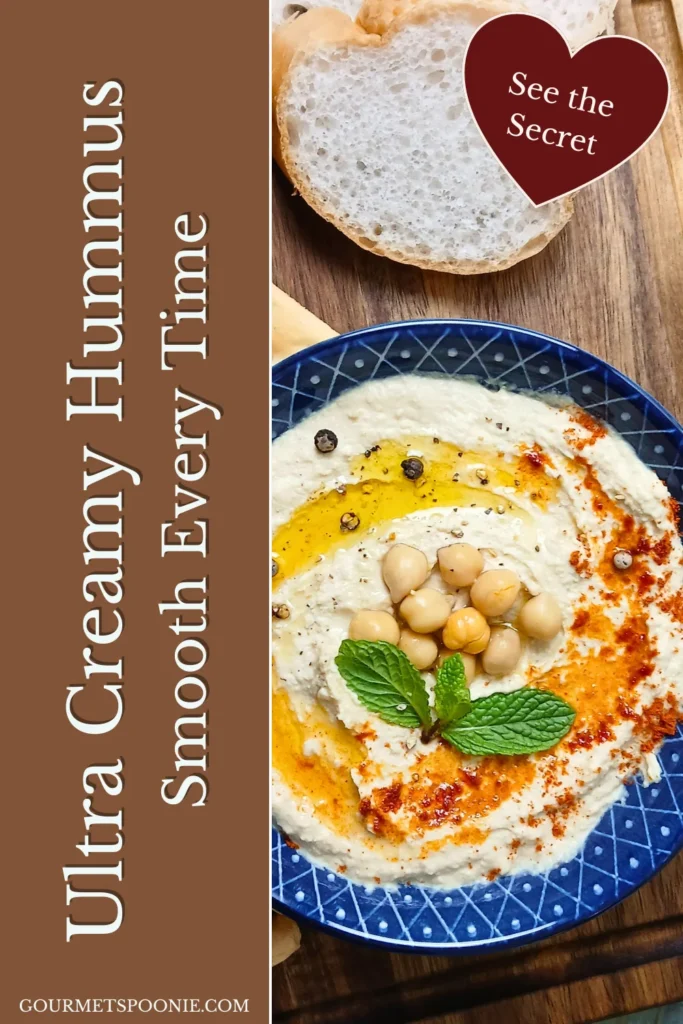 A bowl of ultra creamy hummus garnished with chickpeas, olive oil, spices, and mint sits next to a sliced loaf of bread on a wooden surface.