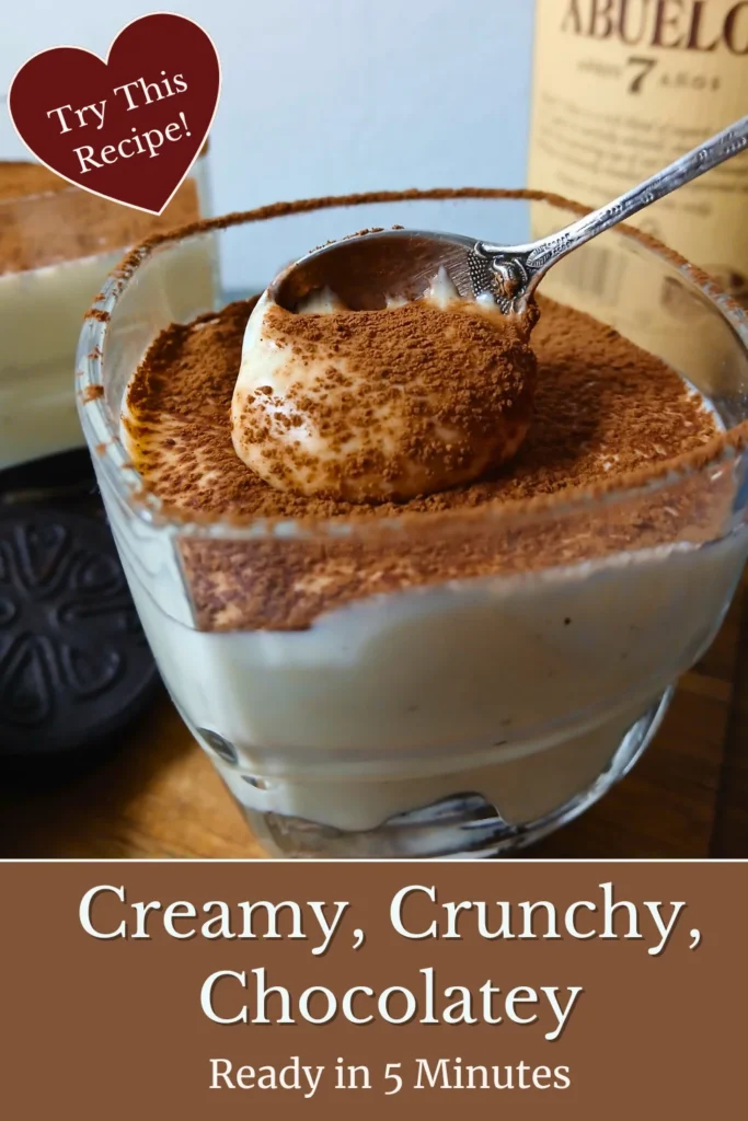 Close-up of a spoon lifting a bite from a vegan tiramisu-style cheesecake cup, revealing layers of vanilla quark and biscuit pieces beneath a cocoa topping.