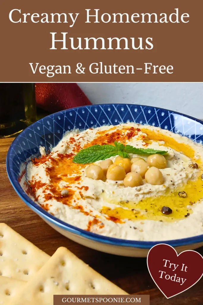 A bowl of creamy homemade hummus topped with olive oil, chickpeas, spices, and a mint leaf, served with crackers on a wooden surface. Text describes it as vegan and gluten-free.