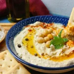 A bowl of easy homemade hummus garnished with chickpeas, olive oil, spices, and mint, surrounded by crackers and sliced bread; a cracker is being dipped into the creamy houmous.