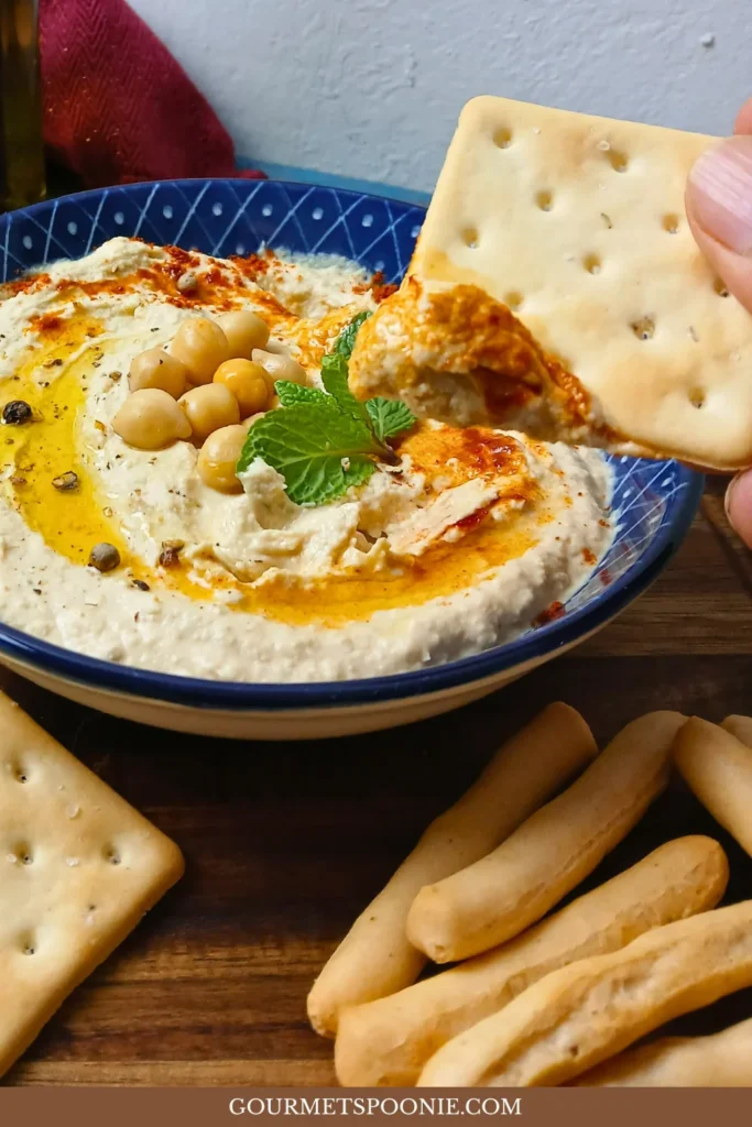 A bowl of easy homemade hummus garnished with chickpeas, olive oil, spices, and mint, surrounded by crackers and sliced bread; a cracker is being dipped into the creamy houmous.