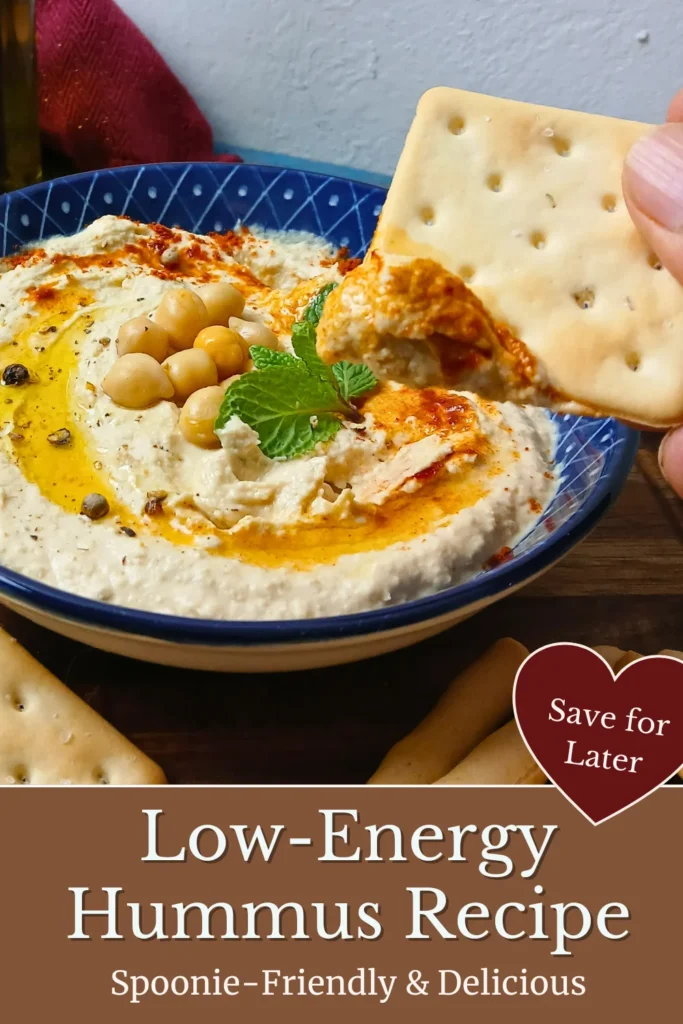 A hand dips a biscuit into a bowl of low-energy houmous recipe, garnished with chickpeas, herbs, olive oil, and spices. Text reads: Low-Energy Hummus Recipe, Spoonie-Friendly and Delicious.