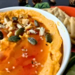 A close-up of a bowl of savoury pumpkin hummus topped with pumpkin seeds, chopped nuts, coarse salt, and a drizzle of sweet chili sauce. A few gluten-free crackers are visible on the side.