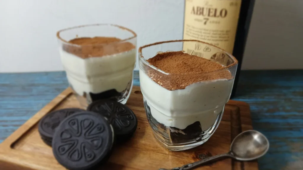 Two vegan tiramisu-style cheesecake cups made with gluten-free Oreo-style biscuits, vanilla quark, and cocoa powder, served on a wooden board beside biscuits and a bottle of dark rum.