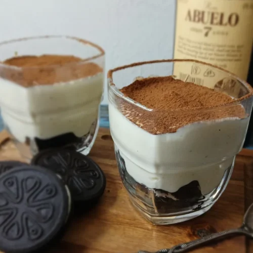 Two vegan tiramisu-style cheesecake cups made with gluten-free Oreo-style biscuits, vanilla quark, and cocoa powder, served on a wooden board beside biscuits and a bottle of dark rum.