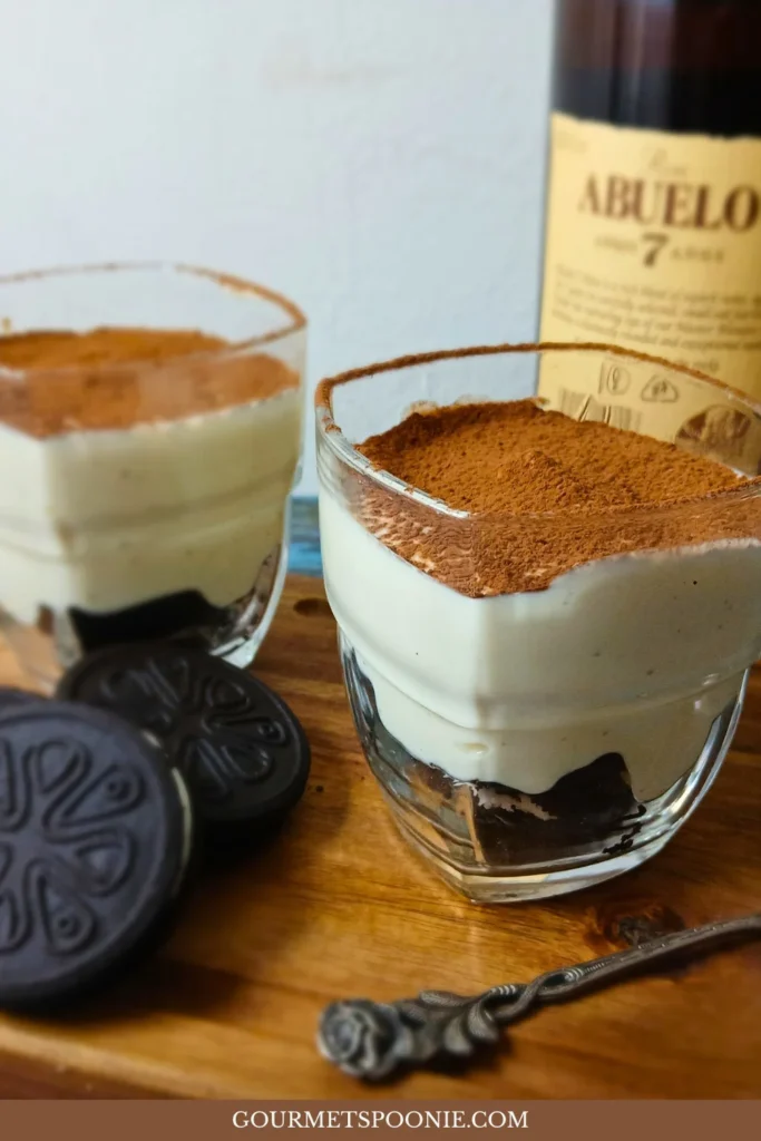 Two vegan tiramisu-style cheesecake cups made with gluten-free Oreo-style biscuits, vanilla quark, and cocoa powder, served on a wooden board beside biscuits and a bottle of dark rum.