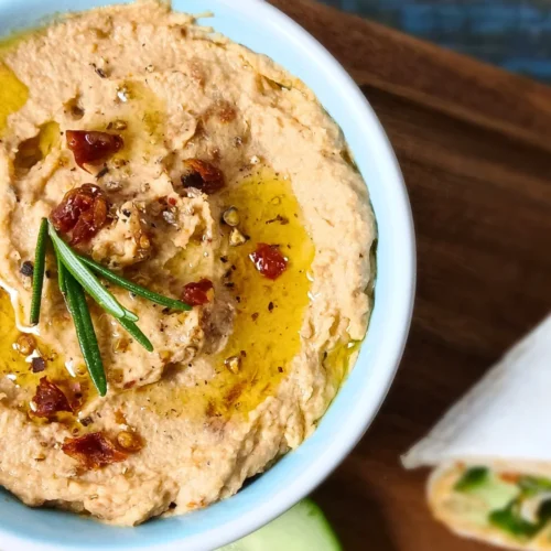 Bowl of creamy sun-dried tomato hummus topped with olive oil and fresh rosemary, served with cucumber and wraps.