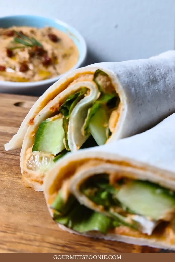 Gluten-free wrap filled with hummus and cucumber beside a bowl of homemade sun-dried tomato hummus.