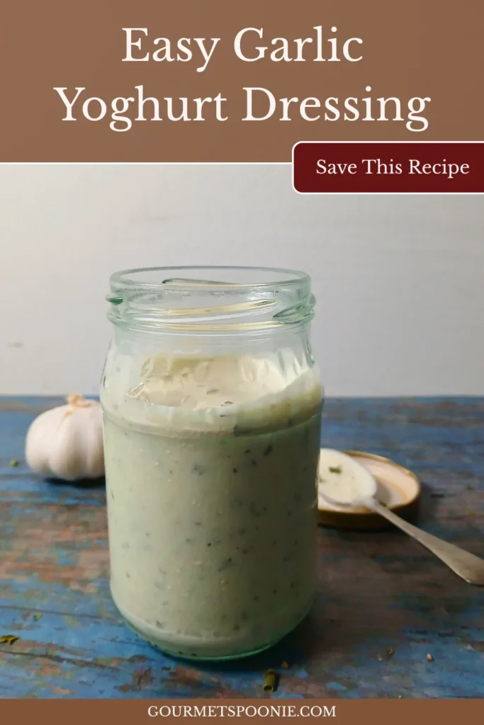 Jar of creamy homemade garlic yoghurt dressing made with Greek yoghurt and herbs, served with garlic cloves and a spoon.