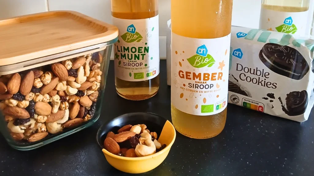 A glass of diluted fruit squash, a bowl of trail mix, gluten-free chocolate biscuits, and a bottle of ginger syrup arranged on a kitchen counter as part of a heatwave preparation plan.