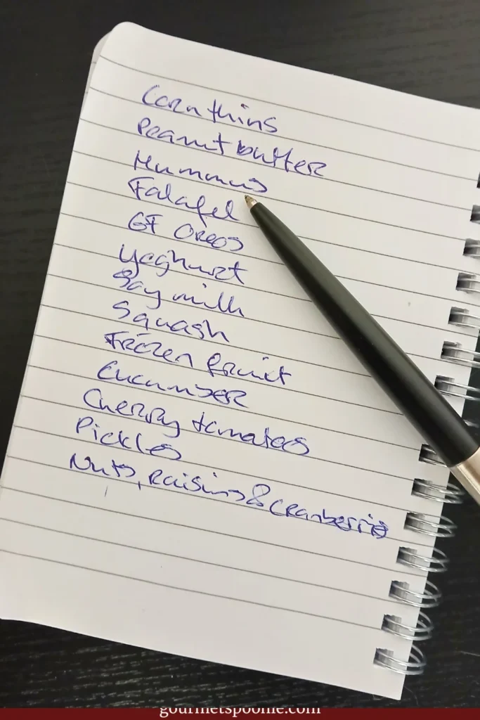 A handwritten shopping list beside a black pen on a notebook, photographed on a dark tabletop, representing planning and shopping for an upcoming heatwave.