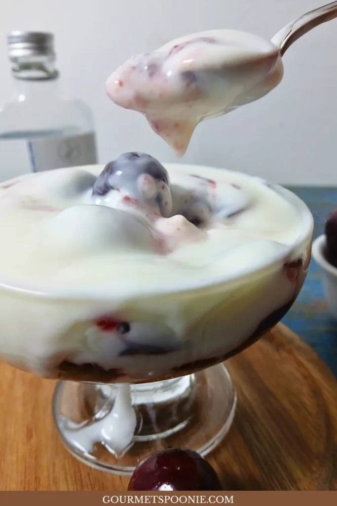 Spoon lifting creamy yoghurt and cherries from a homemade cherry yoghurt trifle in a glass serving bowl.