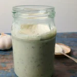 Jar of creamy homemade garlic yoghurt dressing made with Greek yoghurt and herbs, served with garlic cloves and a spoon.