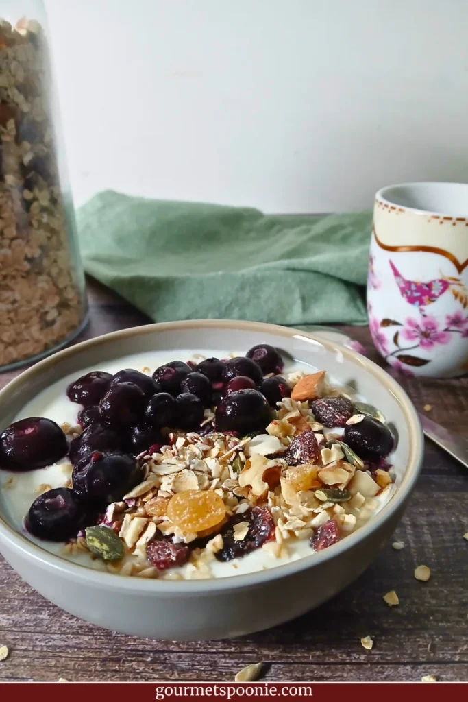 A bowl of vegan yoghurt topped with gluten-free muesli, blueberries, nuts, and seeds beside a mug on a wooden table.